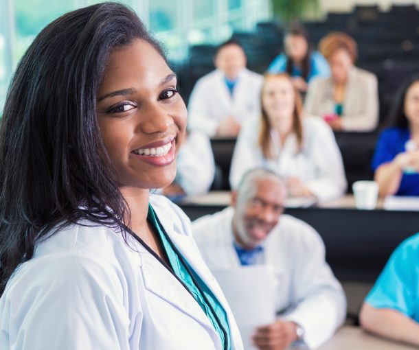 Young adult African American female doctor is standing in hospital lecture hall meeting room. She is attending medical conference or healthcare class with diverse doctors, nurses, and medical students. Adults are wearing scrubs and lab coats.