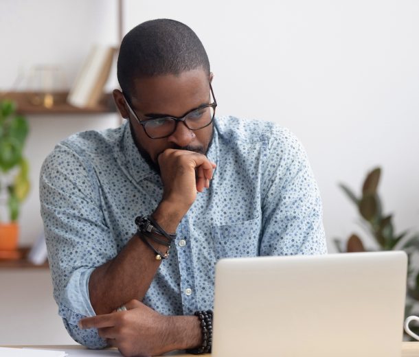 Head shot serious puzzled African American businessman looking at laptop screen sitting in office. Executive managing thinking received bad news keeping fist at chin waiting hoping positive result